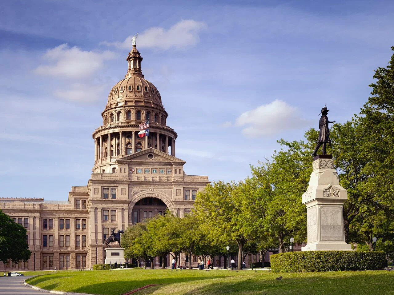 The Texas State Capitol in Austin, its domed tower flying the United States and Texas flags, seen across a lawn with trees and a monument on a tall pedestal.